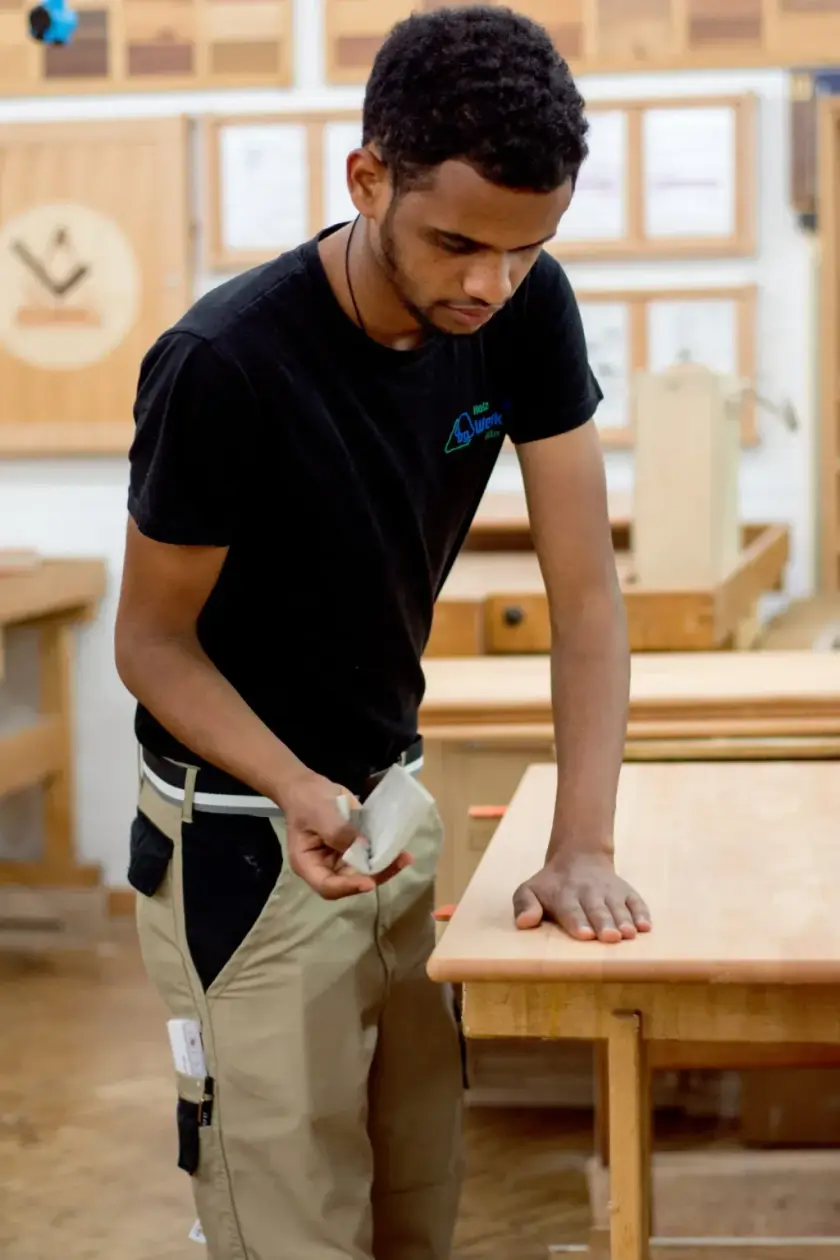 Un jeune homme observe attentivement une planche de bois dans un atelier de menuiserie.