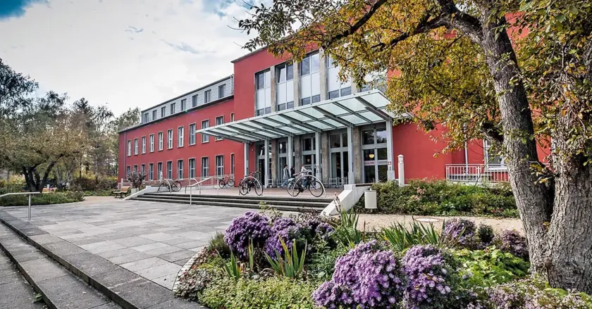 Bâtiment moderne rouge avec une entrée en verre, entouré de fleurs et de verdure.