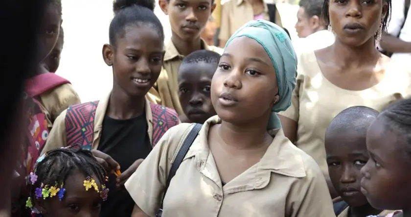 Un groupe d'enfants en uniforme scolaire, certains souriant, d'autres écoutant attentivement une fille au premier plan qui parle.