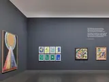 Salle d'exposition avec des œuvres d'art colorées accrochées sur un mur gris.