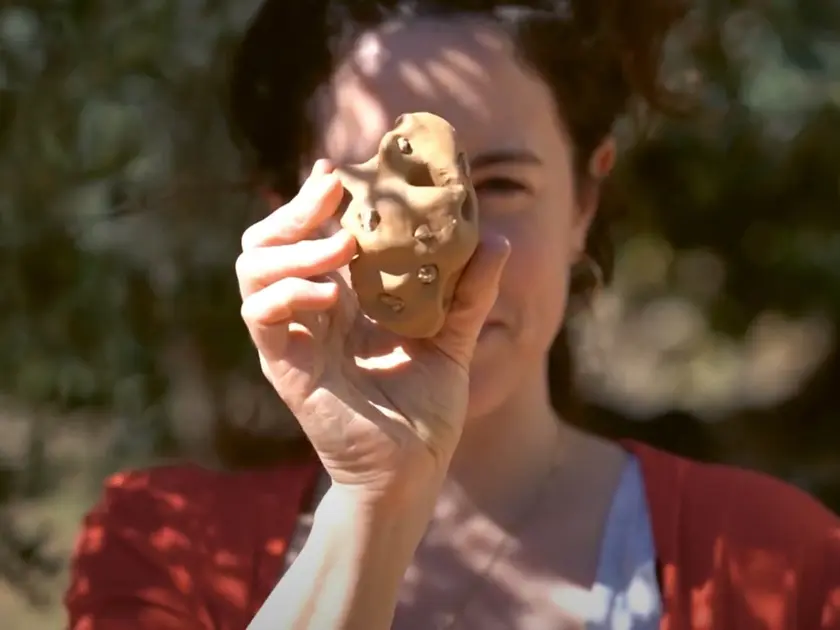 Une femme tient une pomme de terre avec des formes irrégulières, en arrière-plan flou d'une nature verdoyante.