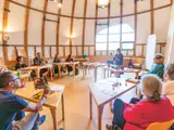 Un groupe de personnes assises autour de tables dans une salle lumineuse, écoutant une intervenante.