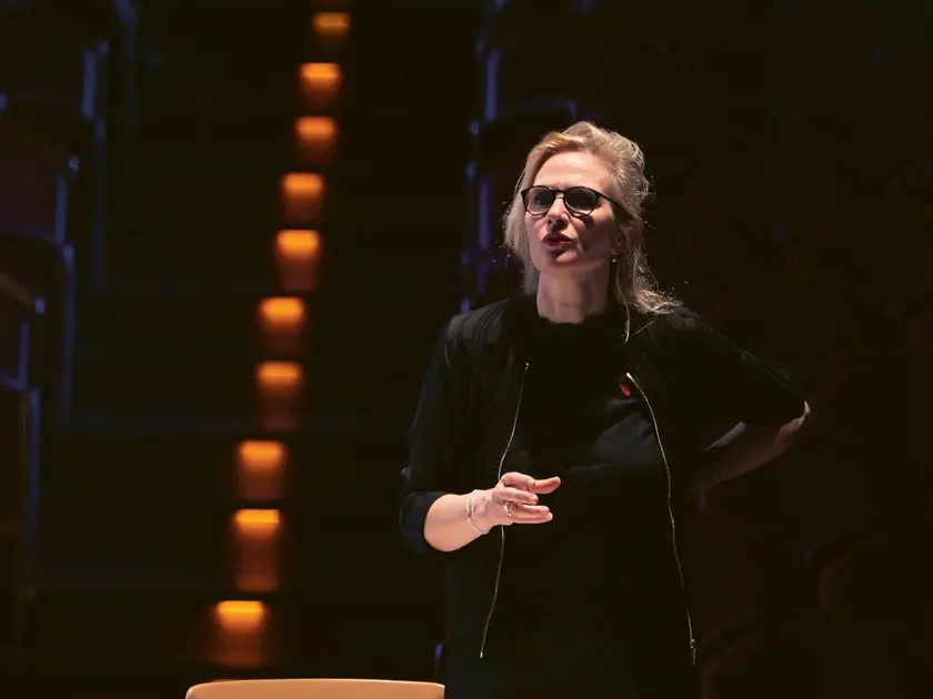 Femme en lunettes parlant sur scène, avec des sièges en arrière-plan et un éclairage tamisé.