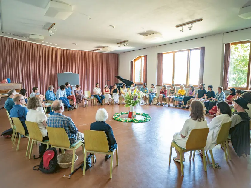 Groupe de personnes assises en cercle dans une salle lumineuse, avec un arrangement floral au centre.