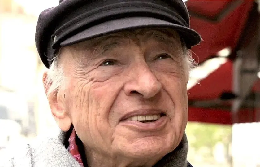 Un homme âgé souriant, portant une casquette noire et un manteau, avec un fond flou.