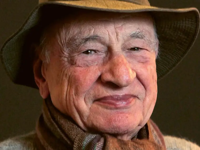 Un homme âgé souriant, portant un chapeau et une écharpe, avec un fond sombre.