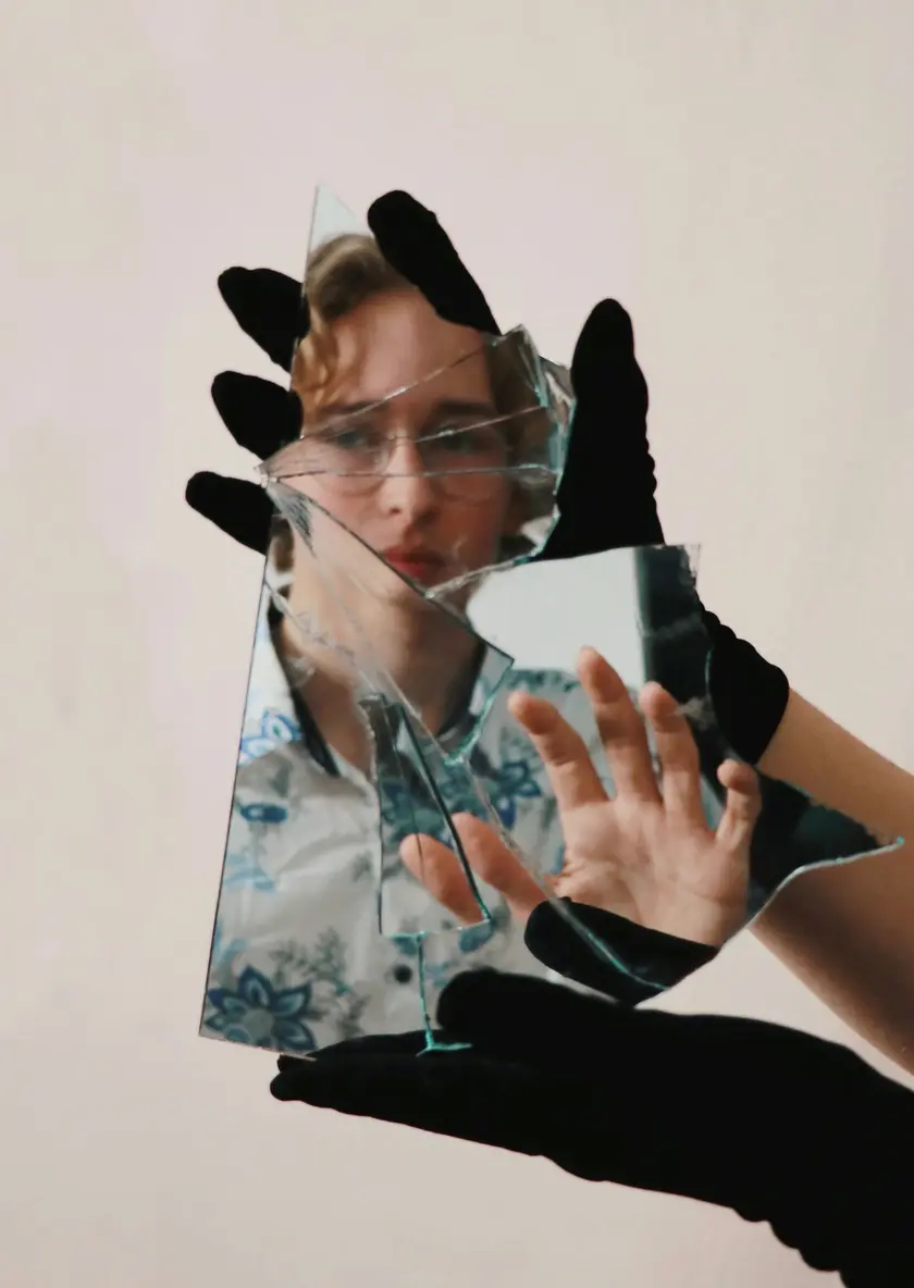 Une main gantée tient un miroir brisé, reflétant un visage en arrière-plan.