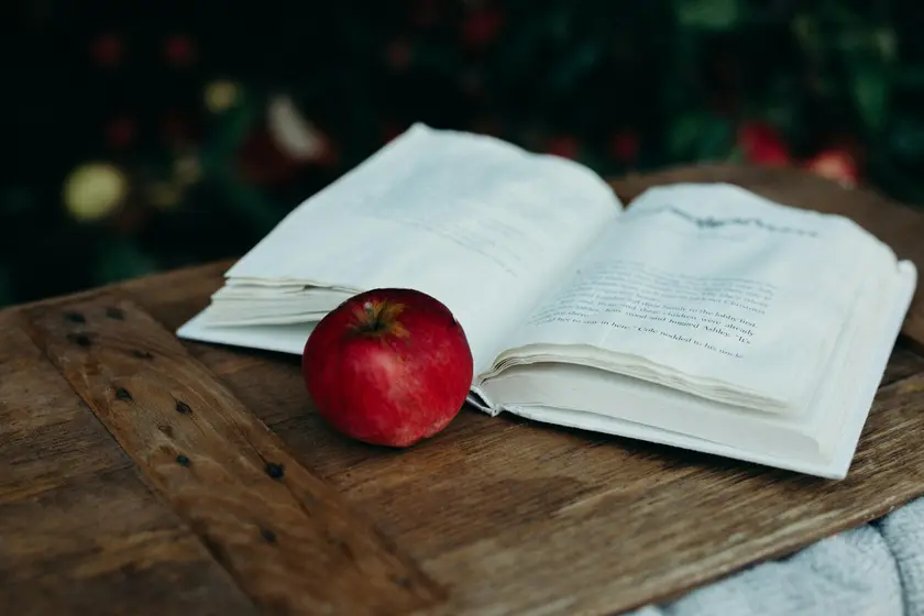 Un livre ouvert posé sur une table en bois, avec une pomme rouge à côté.