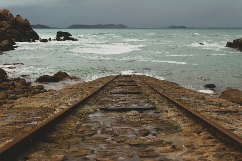 Des rails de train menant vers la mer, entourés de rochers et d'une mer agitée sous un ciel nuageux.