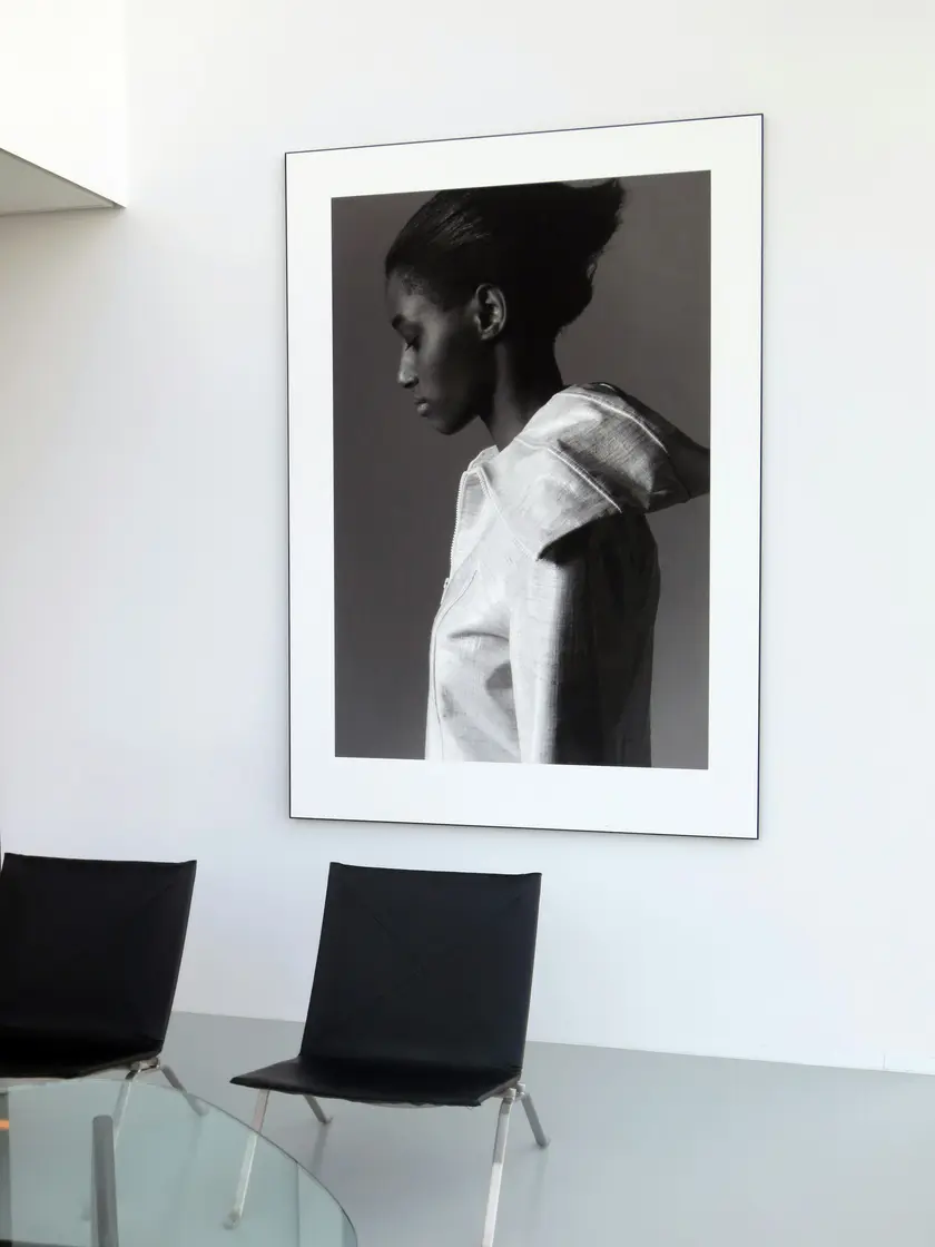 Portrait en noir et blanc d'une femme de profil, affiché sur un mur blanc, avec des chaises noires en avant-plan.